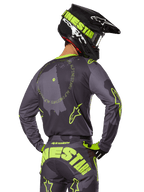 Piloto de motocross visto desde atrás con el conjunto de equipamiento Racer Hollow 2025, jersey gris con texto circular y logotipos de estrellas en amarillo fluorescente, pantalones grises y amarillos a juego con marca llamativa en el asiento, casco negro y verde lima, atuendo coordinado para carreras fuera de carretera
