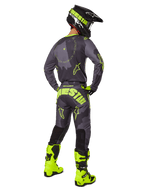 Vista posterior de un piloto de motocross que viste el conjunto Racer Hollow 2025 en color Dark Camo Yellow Fluo, con un jersey de camuflaje gris con detalles en amarillo flúor, pantalones de motocross a juego con un logotipo llamativo en el asiento, botas amarillo flúor vibrante y un casco negro con gafas amarillas.