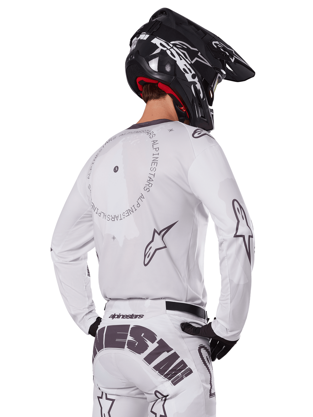 Piloto de motocross desde atrás con el conjunto Alpinestars 2025 Racer Hollow en color blanco, jersey blanco de manga larga con logotipos de estrellas en gris y gráficos circulares, pantalones blancos a juego con marca llamativa en el asiento, casco negro, equipamiento de carreras todoterreno coordinado.