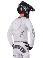 Piloto de motocross desde atrás con el conjunto Alpinestars 2025 Racer Hollow en color blanco, jersey blanco de manga larga con logotipos de estrellas en gris y gráficos circulares, pantalones blancos a juego con marca llamativa en el asiento, casco negro, equipamiento de carreras todoterreno coordinado.
