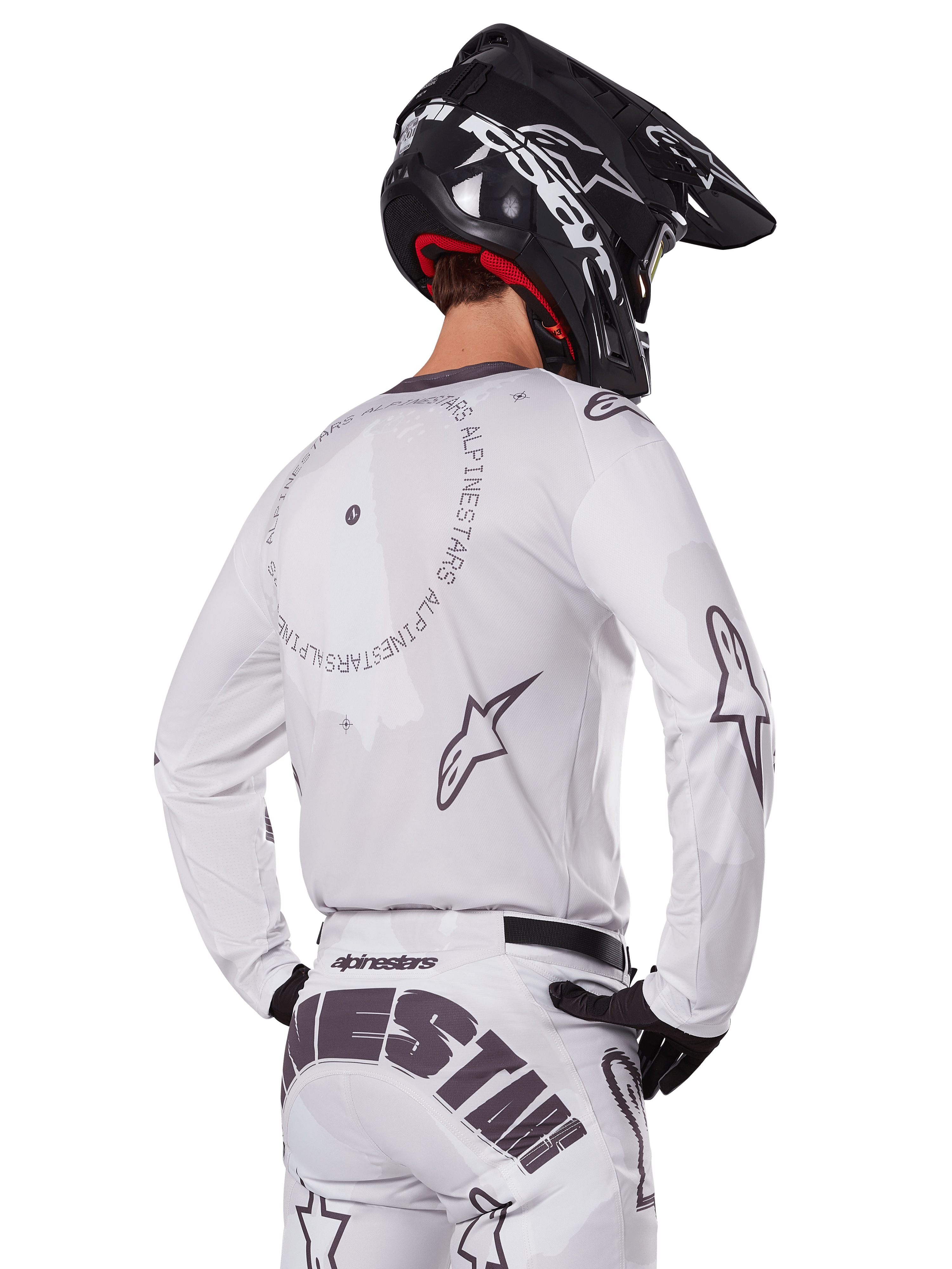 Piloto de motocross desde atrás con el conjunto Alpinestars 2025 Racer Hollow en color blanco, jersey blanco de manga larga con logotipos de estrellas en gris y gráficos circulares, pantalones blancos a juego con marca llamativa en el asiento, casco negro, equipamiento de carreras todoterreno coordinado.