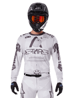 Alpinestars 2025 Racer Hollow Jersey, camiseta de motocross, blanca y negra, usada con pantalones de MX blancos a juego y un casco negro con gafas naranjas, con marca Alpinestars destacada y gráficos de globo, equipo profesional de carreras todoterreno