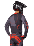 Alpinestars 2025 Racer Hollow Jersey, vista trasera de un piloto de motocross con equipamiento en Gris Camo y Naranja Flúor, con pantalones de camuflaje a juego con letras grandes de Alpinestars en naranja y un casco negro, diseño de ropa profesional para off-road.