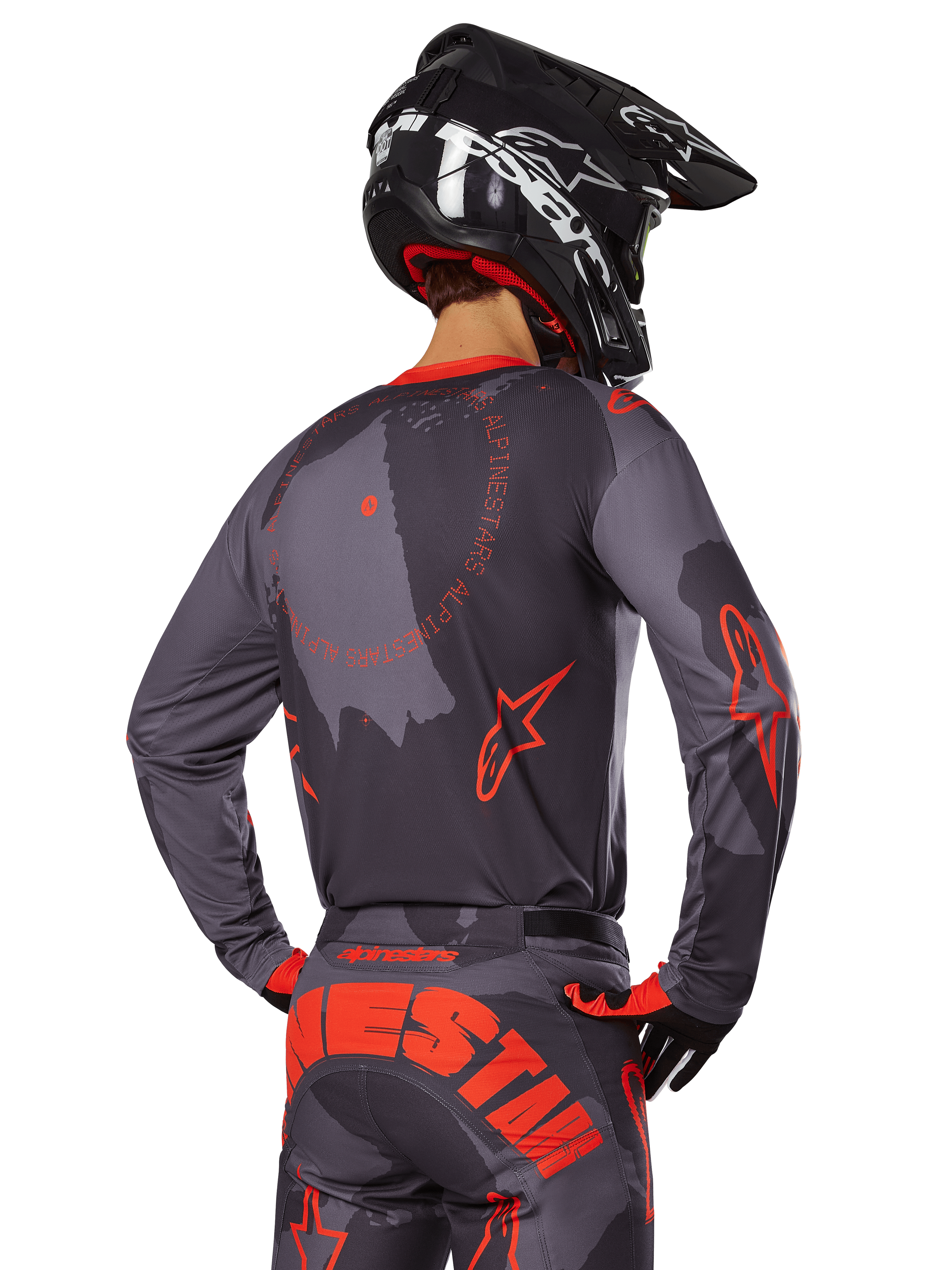 Alpinestars 2025 Racer Hollow Jersey, vista trasera de un piloto de motocross con equipamiento en Gris Camo y Naranja Flúor, con pantalones de camuflaje a juego con letras grandes de Alpinestars en naranja y un casco negro, diseño de ropa profesional para off-road.