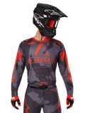 Alpinestars 2025 Racer Hollow Jersey, jersey de motocross, gris camuflaje y naranja flúor, combinado con un conjunto a juego de pantalones y guantes, con casco negro con gráficos blancos y gafas, logotipo ASTARS destacado, ropa deportiva protectora para todoterreno