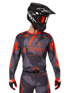 Alpinestars 2025 Racer Hollow Jersey, jersey de motocross, gris camuflaje y naranja flúor, combinado con un conjunto a juego de pantalones y guantes, con casco negro con gráficos blancos y gafas, logotipo ASTARS destacado, ropa deportiva protectora para todoterreno