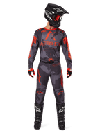 Piloto de motocross con jersey Alpinestars 2025 Racer Hollow y pantalones a juego en Gris Camo Naranja Fluo, conjunto de equipamiento completo con casco negro, gafas, guantes y botas, con logotipos naranjas llamativos y estampado de camuflaje.