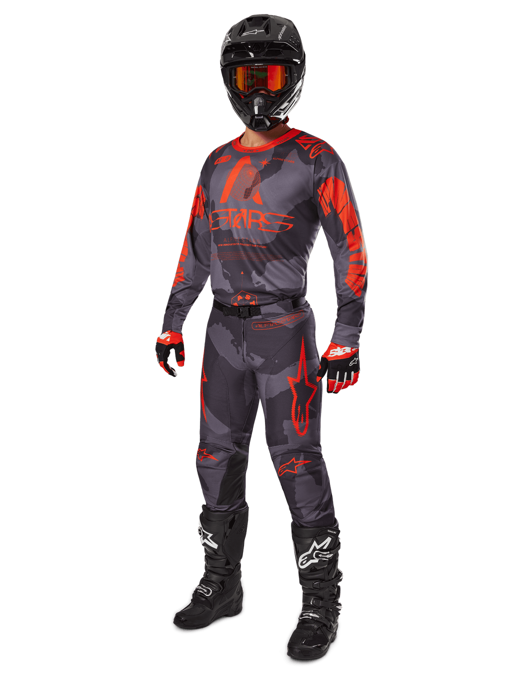 Piloto de motocross con equipamiento completo de competición, jersey y pantalones Alpinestars 2025 Racer Hollow en Gris Camo Naranja Flúor, casco negro con gafas naranjas, guantes negros y rojos, y botas negras con logotipos blancos, conjunto de alto rendimiento coordinado para carreras off-road