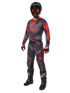 Piloto de motocross con equipamiento completo de competición, jersey y pantalones Alpinestars 2025 Racer Hollow en Gris Camo Naranja Flúor, casco negro con gafas naranjas, guantes negros y rojos, y botas negras con logotipos blancos, conjunto de alto rendimiento coordinado para carreras off-road