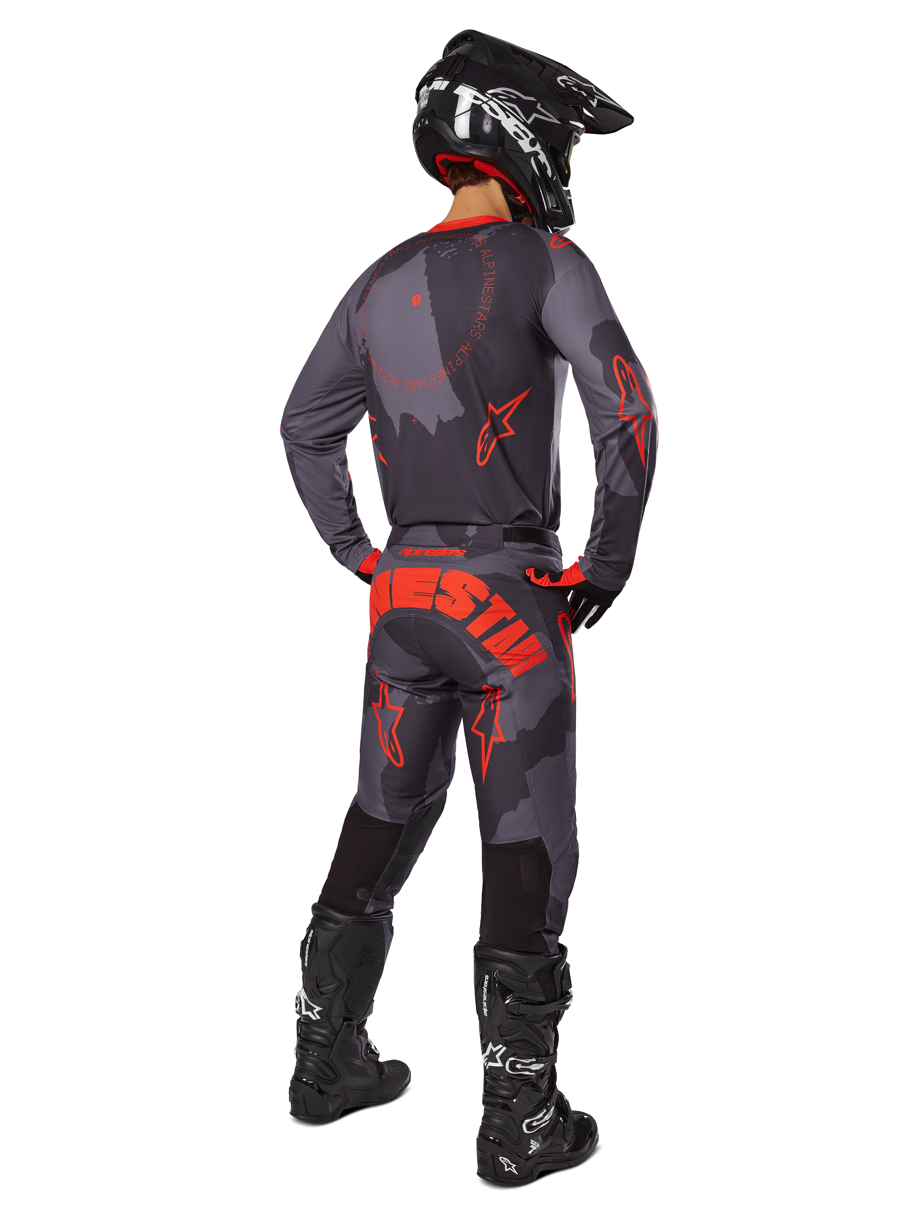 Piloto de motocross visto desde atrás con el jersey Alpinestars 2025 Racer Hollow en gris camuflaje y naranja flúor, pantalones de camuflaje a juego con texto de Alpinestars en naranja, casco negro y botas negras, conjunto de equipamiento coordinado para conducción off-road