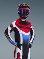 Alpinestars 2026 Techstar Knif Gear Set, piloto de motocross con jersey de carreras rojo, blanco y azul con detalles negros, casco y gafas multicolores a juego, guantes negros, equipo profesional para competición de motocross.