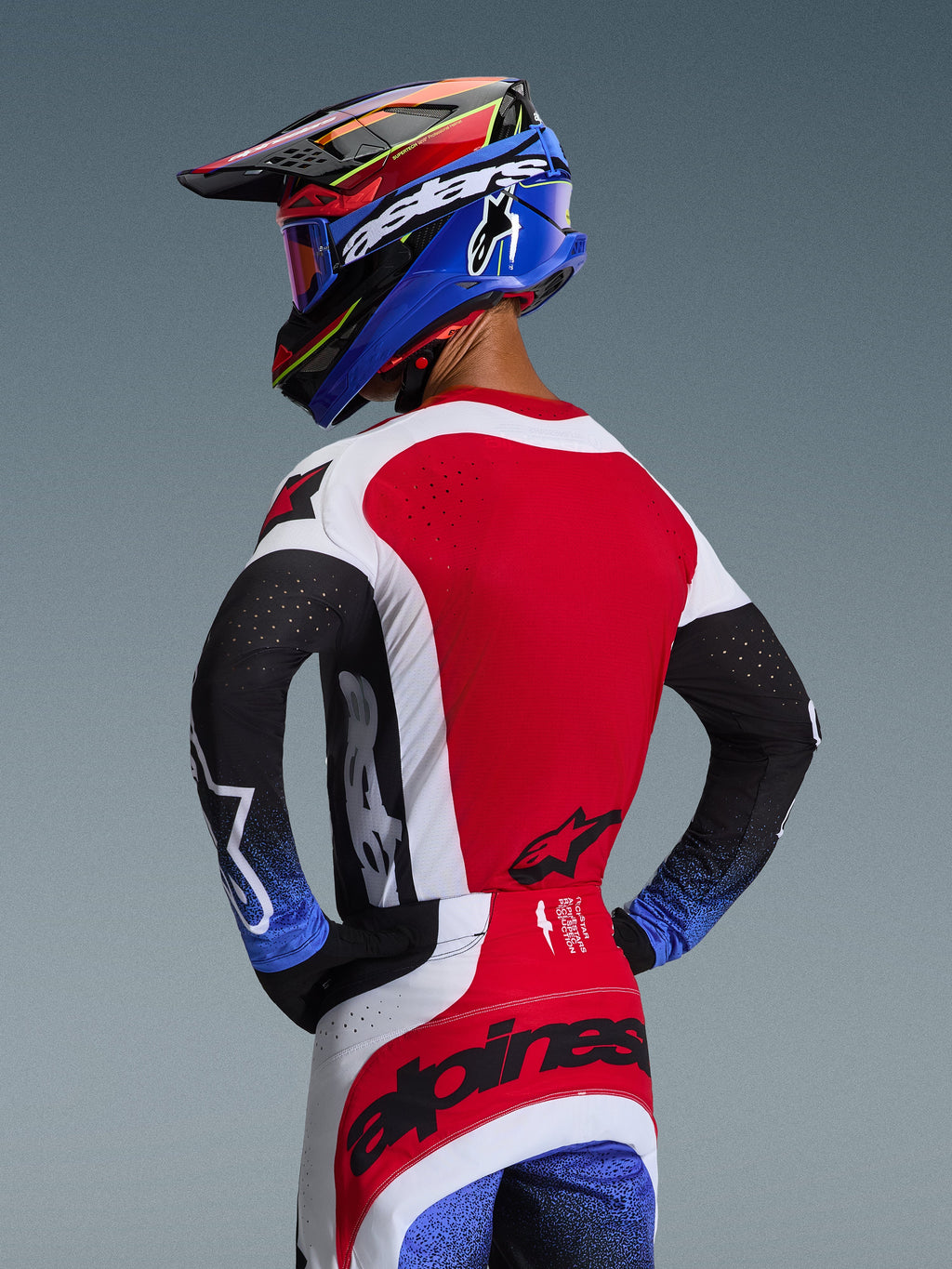 Piloto de motocross visto desde atrás con el conjunto Techstar Knif 2026, jersey rojo con paneles laterales blancos y mangas negras con puños moteados en azul, pantalones rojos y blancos con la marca Alpinestars en negro en el asiento, casco azul y rojo a juego con gafas