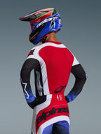 Piloto de motocross visto desde atrás con el conjunto Techstar Knif 2026, jersey rojo con paneles laterales blancos y mangas negras con puños moteados en azul, pantalones rojos y blancos con la marca Alpinestars en negro en el asiento, casco azul y rojo a juego con gafas
