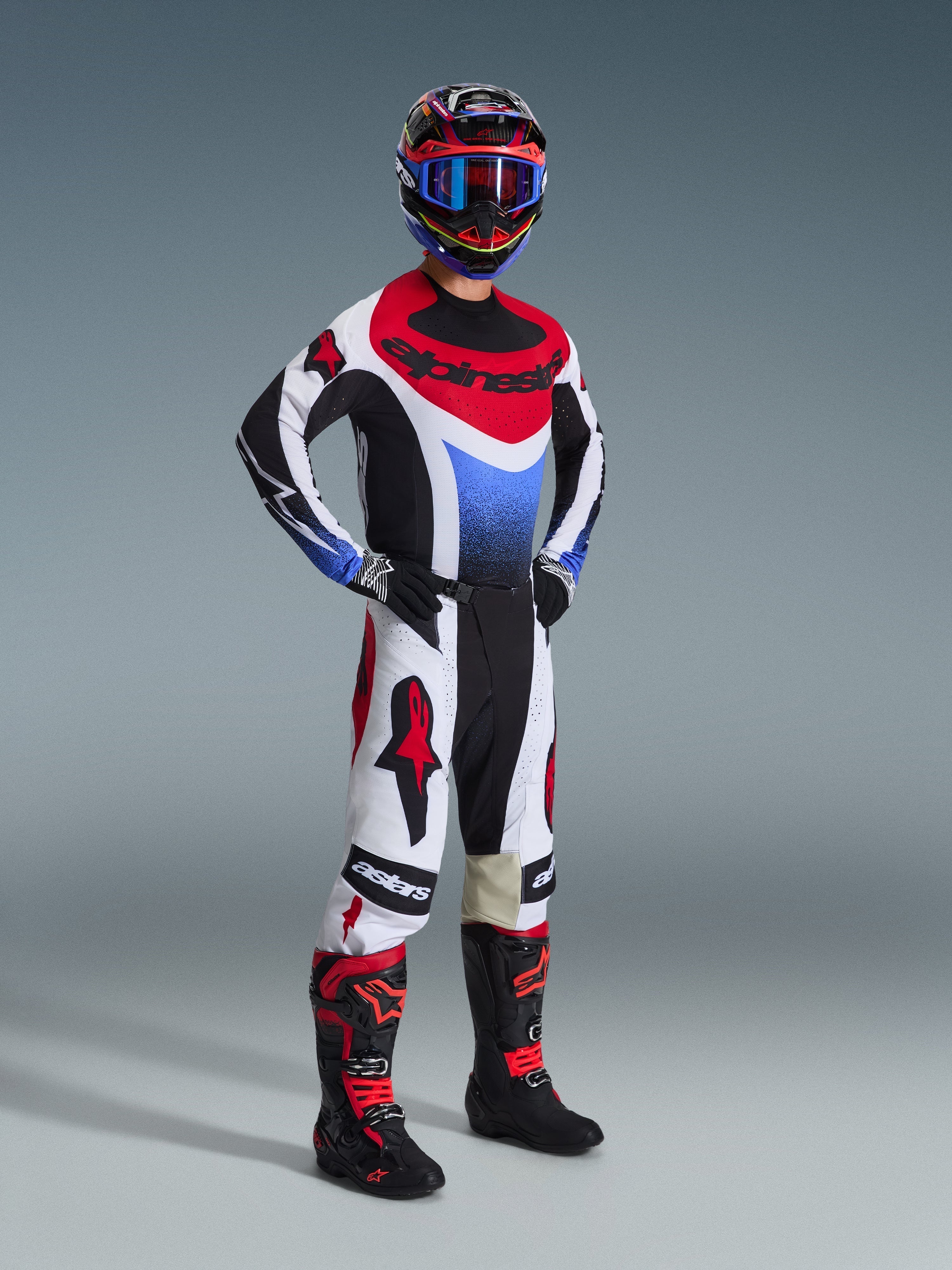 Piloto de motocross con el conjunto de equipación Alpinestars Techstar Knif 2026, jersey con paneles en negro, blanco y rojo con un degradado azul en el pecho, pantalones blancos con logotipos rojos de Alpinestars y detalles negros, botas negras y rojas; conjunto profesional completo para competición off-road.