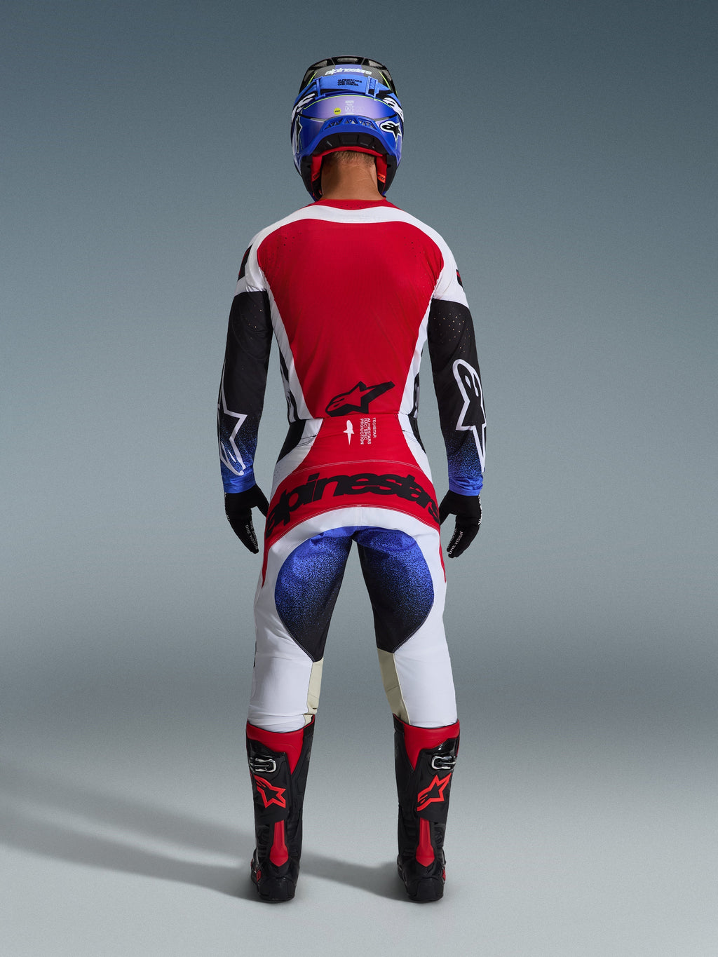 Piloto de motocross con el conjunto Alpinestars Techstar Knif 2026 visto desde atrás, con un jersey rojo, blanco y negro con detalles en degradado azul en las mangas, pantalones blancos y negros a juego con logotipos rojos llamativos, casco azul y botas negras y rojas, equipo profesional de competición todoterreno.