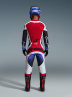 Piloto de motocross con el conjunto Alpinestars Techstar Knif 2026 visto desde atrás, con un jersey rojo, blanco y negro con detalles en degradado azul en las mangas, pantalones blancos y negros a juego con logotipos rojos llamativos, casco azul y botas negras y rojas, equipo profesional de competición todoterreno.
