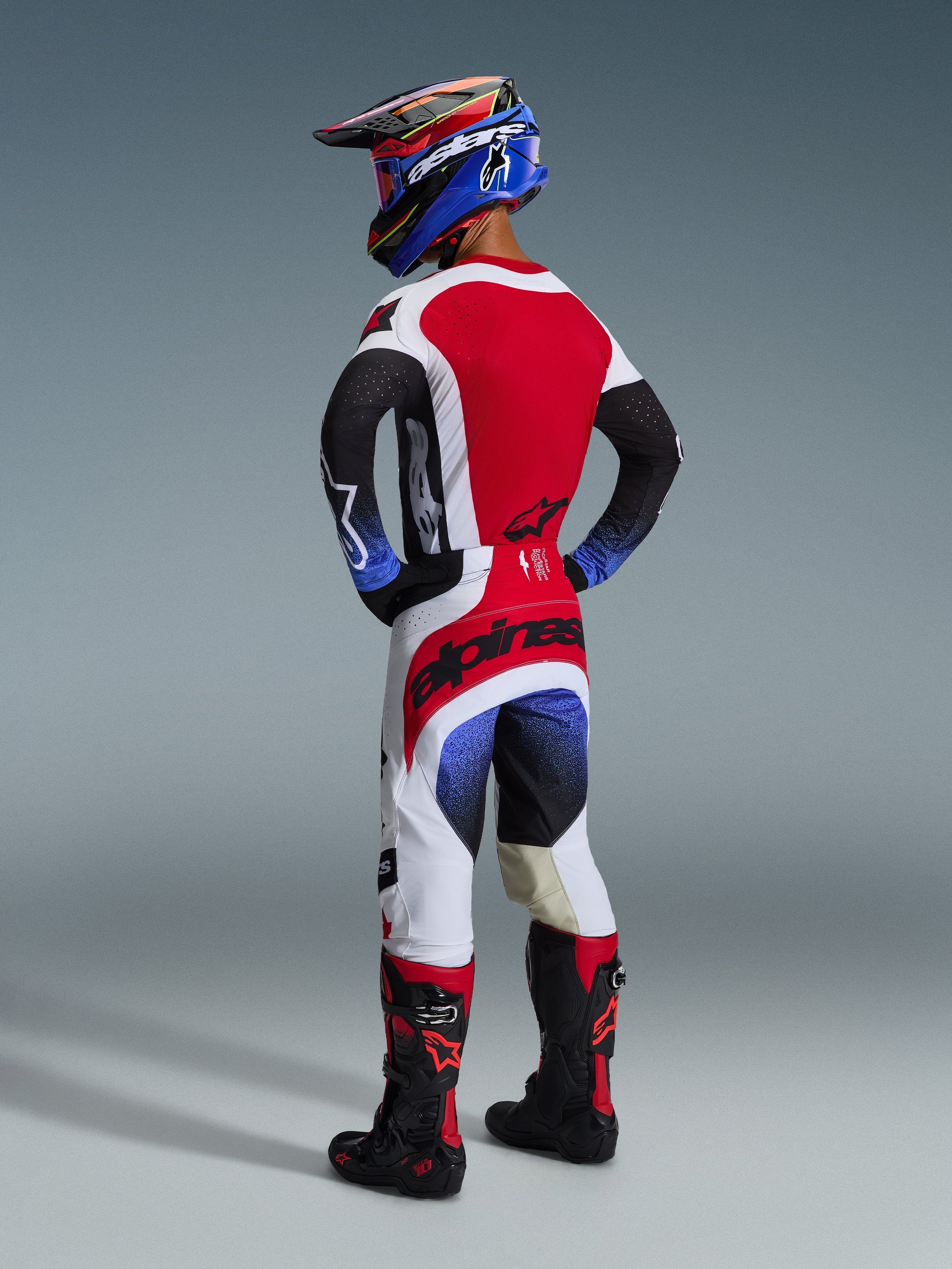Piloto de motocross en vista trasera con el conjunto Techstar Knif 2026, jersey negro, blanco y rojo con mangas en degradado azul y perforaciones cortadas con láser, pantalones blancos y rojos a juego con la marca Alpinestars en negro en el asiento, casco azul y negro con gafas rojas, botas de motocross negras y rojas, kit completo de competición off-road