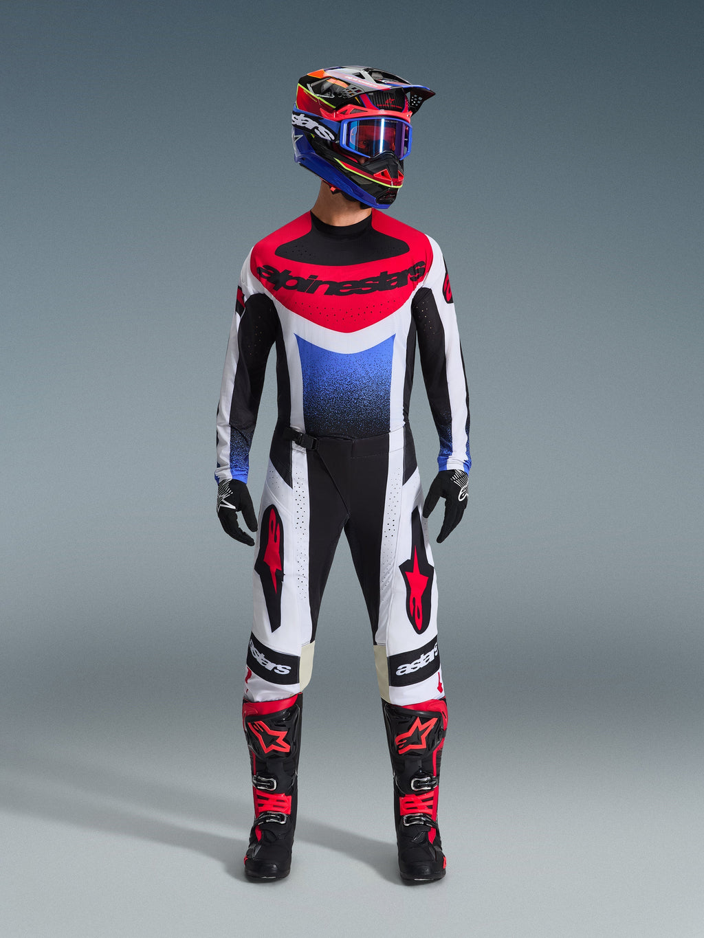 Piloto de motocross con el conjunto de equipación Alpinestars 2026 Techstar Knif, jersey negro, blanco y rojo con degradado azul y logotipos, pantalones a juego en blanco y negro con detalles rojos, casco multicolor con gafas azules y botas negras y rojas, conjunto de competición todoterreno coordinado.