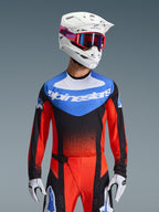 Alpinestars 2026 Techstar Knif Jersey, jersey de motocross, naranja, negro y azul UCLA, con un panel azul en el pecho con logo de Alpinestars en blanco y mangas naranja rojizo, usado con casco blanco, gafas rojas y guantes blancos, equipamiento profesional de motocross