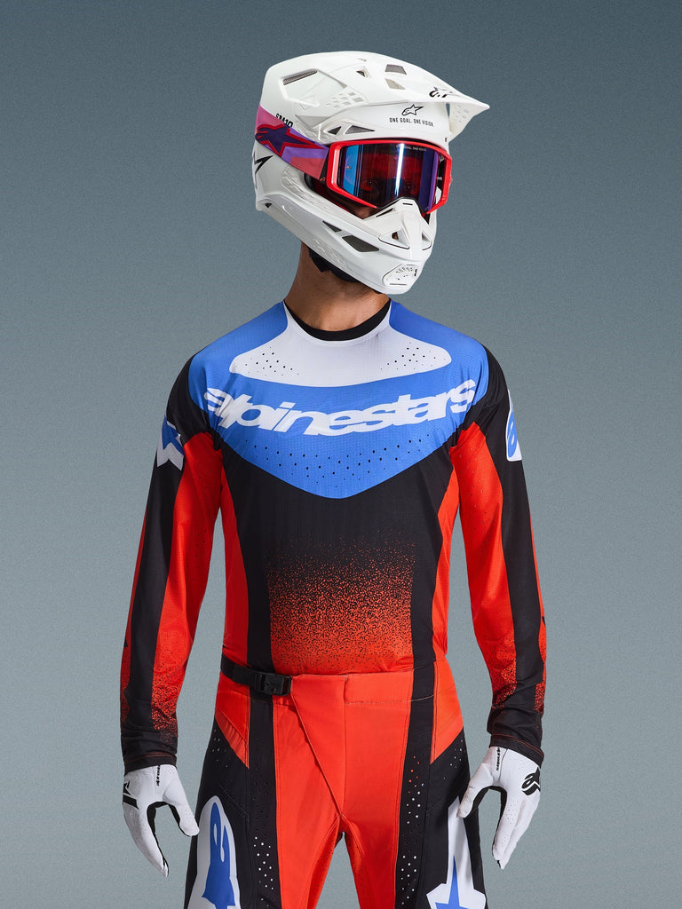 Alpinestars 2026 Techstar Knif Jersey, jersey de motocross, naranja, negro y azul UCLA, con un panel azul en el pecho con logo de Alpinestars en blanco y mangas naranja rojizo, usado con casco blanco, gafas rojas y guantes blancos, equipamiento profesional de motocross