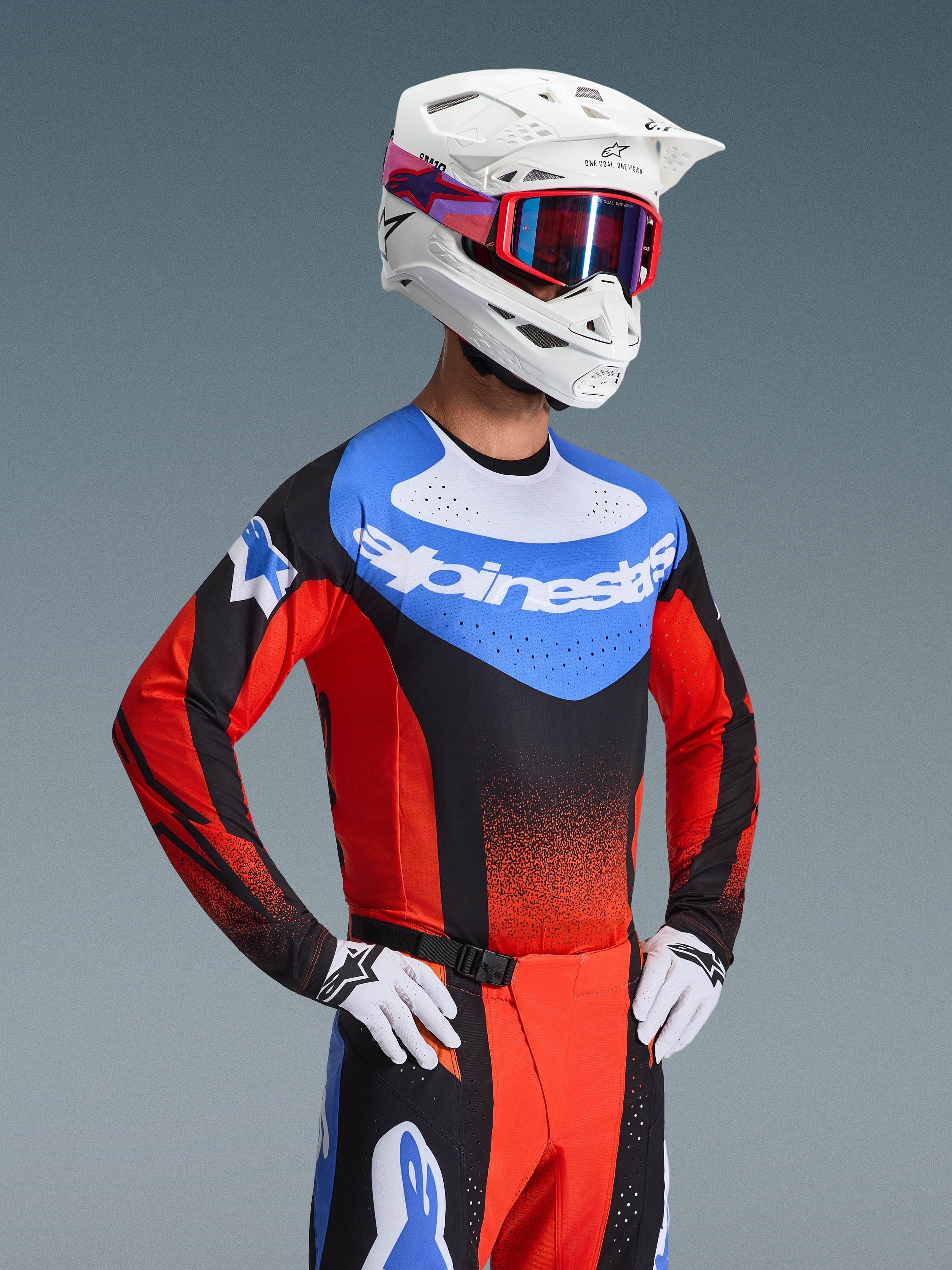 Piloto de motocross con el Jersey Alpinestars Techstar Knif 2026 en naranja, negro y azul UCLA, con un panel azul en el pecho, logo de Alpinestars en blanco y mangas con perforaciones láser, combinado con casco blanco, gafas rojas y pantalones de carreras a juego.