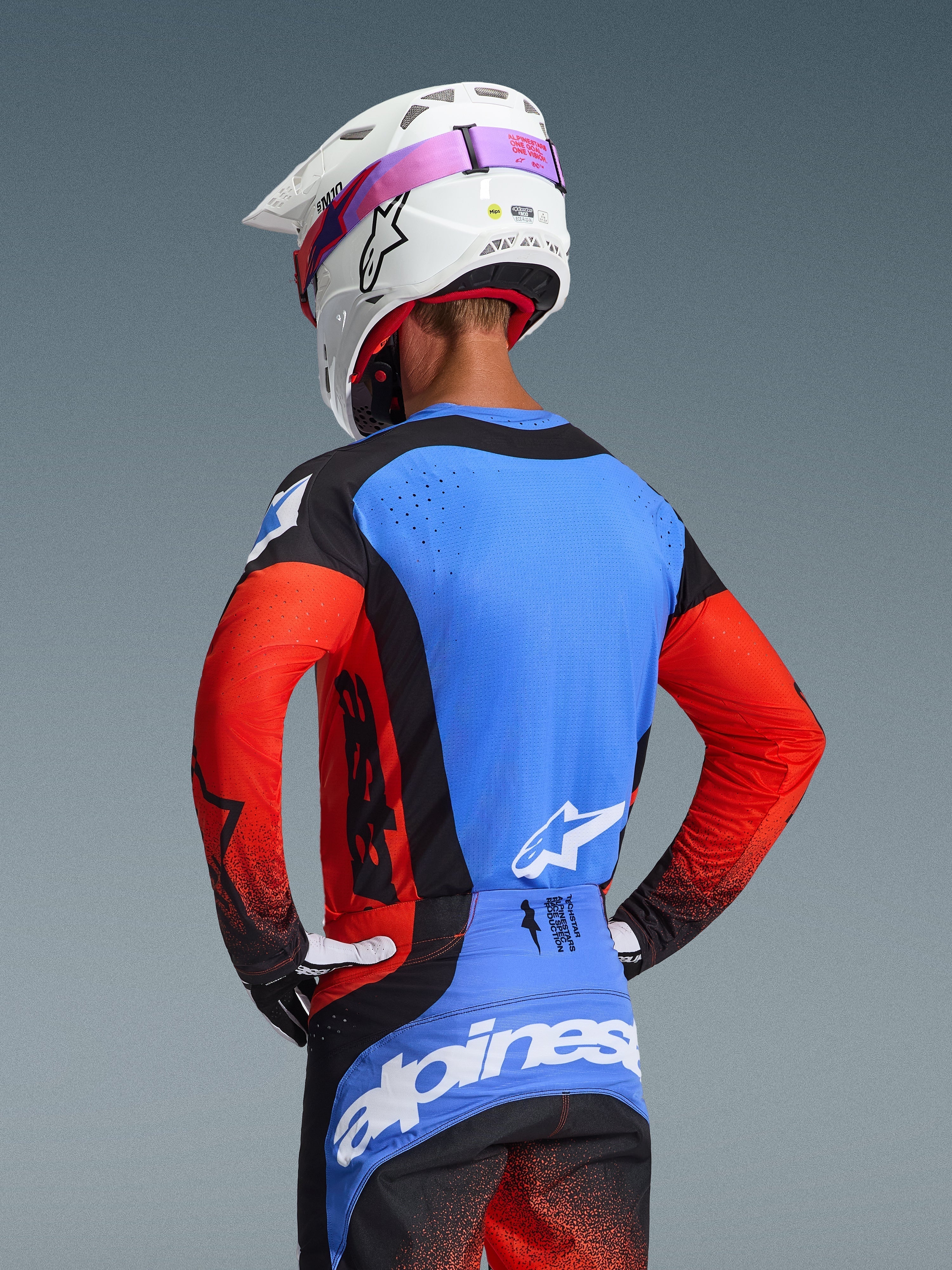 Vista trasera de un piloto de motocross con el Jersey Techstar Knif 2026 en naranja, negro y azul UCLA, con un panel trasero azul con perforaciones cortadas por láser, mangas naranjas y detalles en negro, combinado con pantalones de motocross a juego y un casco Alpinestars blanco.