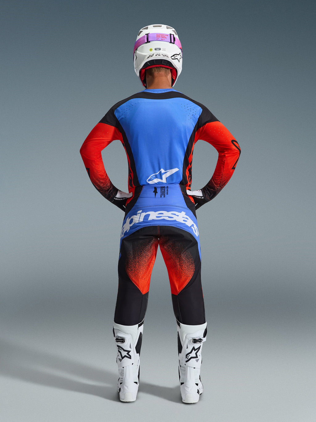 Piloto de motocross visto desde atrás con equipamiento de competición completo, Jersey Techstar Knif 2026 con espalda azul y mangas naranjas, pantalones negros con detalles en naranja y azul, casco blanco con cinta de gafas rosa, botas blancas y negras, conjunto coordinado para carreras off-road
