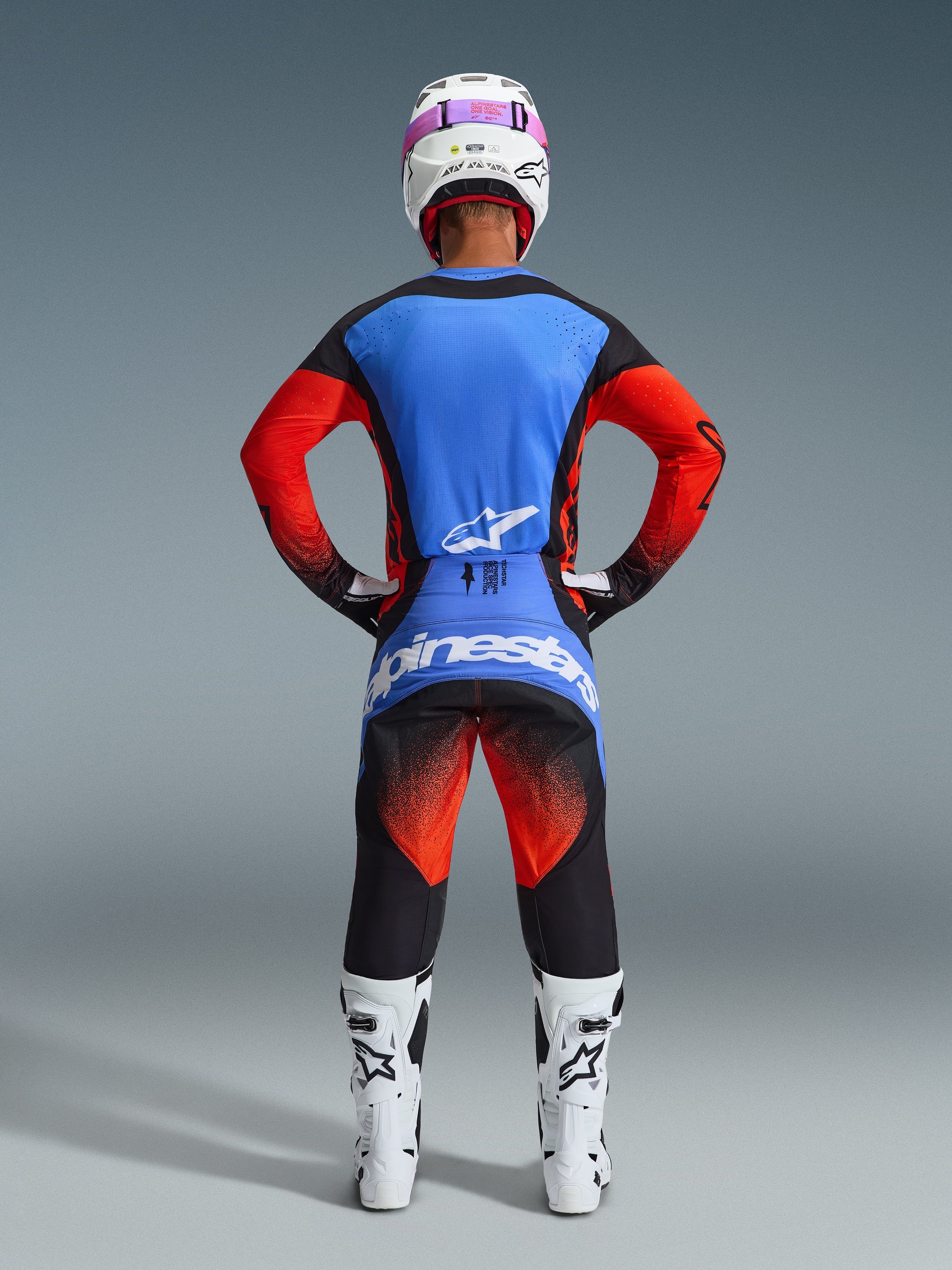 Piloto de motocross visto desde atrás con equipamiento de competición completo, Jersey Techstar Knif 2026 con espalda azul y mangas naranjas, pantalones negros con detalles en naranja y azul, casco blanco con cinta de gafas rosa, botas blancas y negras, conjunto coordinado para carreras off-road