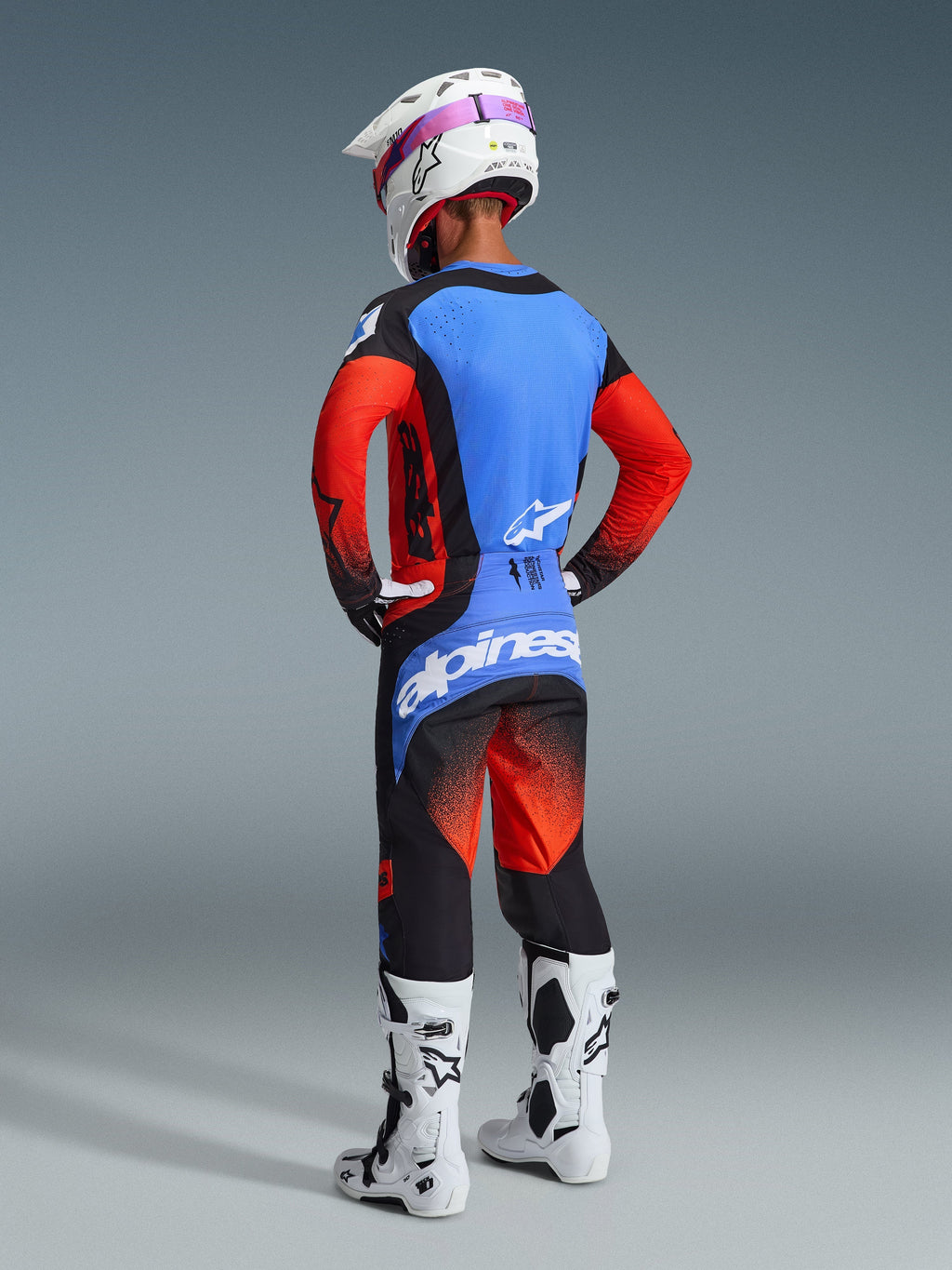 Piloto de motocross visto desde atrás con el Jersey Techstar Knif 2026 en naranja, negro y azul UCLA, combinado con pantalones de motocross a juego, casco blanco con gafas rosas y botas blancas, mostrando un equipamiento completo de Alpinestars para competición off-road.