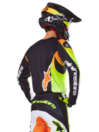 Piloto de motocross visto desde atrás con el conjunto de equipo Alpinestars 2025 Fluid Wurx, jersey negro con gráficos vibrantes en amarillo neón y naranja, pantalones a juego con paneles blancos y naranjas, casco negro con detalles neón, logotipo destacado de Alpinestars en las mangas y el asiento