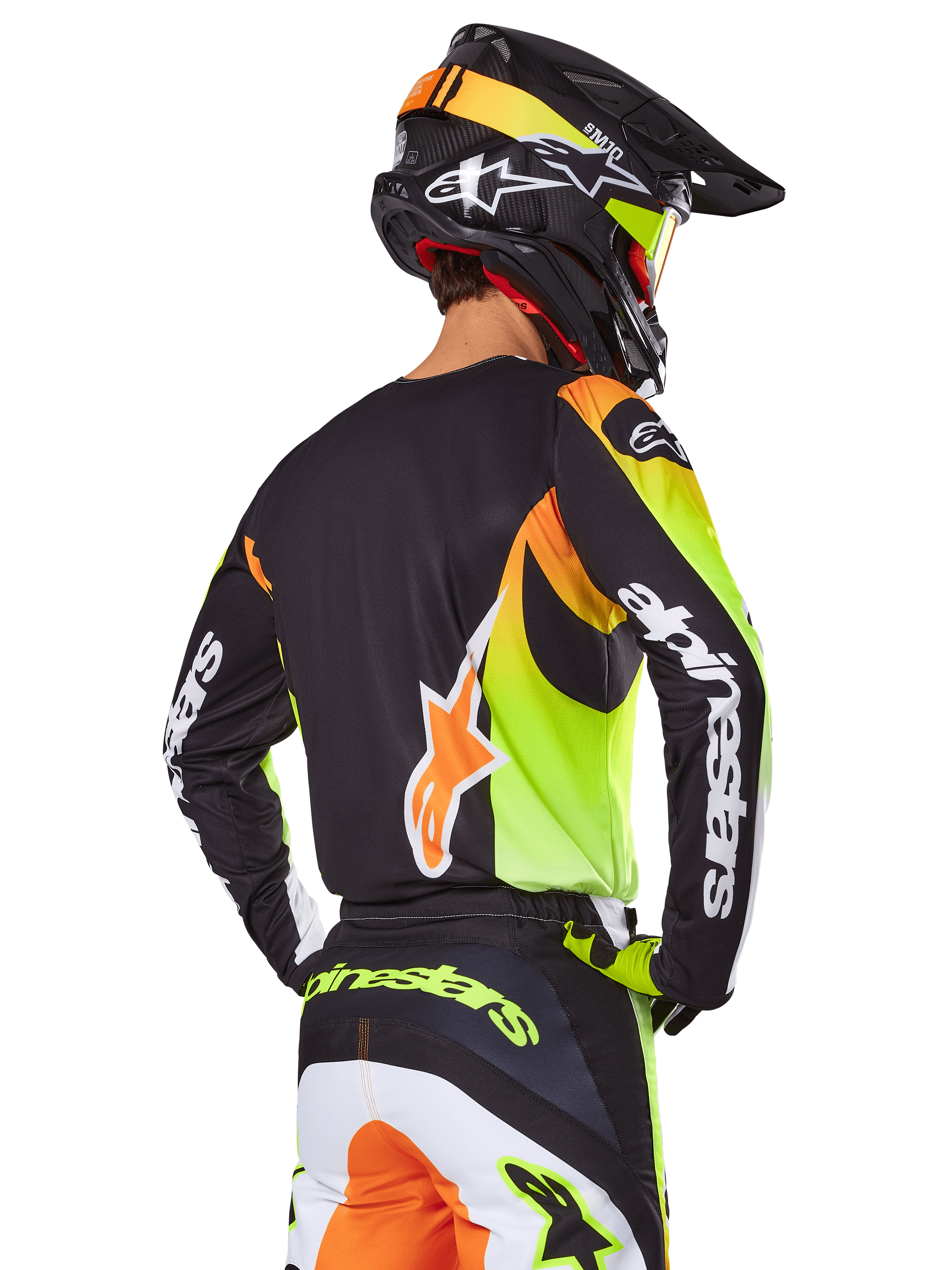 Piloto de motocross visto desde atrás con el conjunto de equipo Alpinestars 2025 Fluid Wurx, jersey negro con gráficos vibrantes en amarillo neón y naranja, pantalones a juego con paneles blancos y naranjas, casco negro con detalles neón, logotipo destacado de Alpinestars en las mangas y el asiento