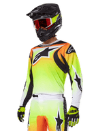Piloto de motocross con el conjunto de equipamiento Alpinestars 2025 Fluid Wurx, jersey con degradado en amarillo y naranja con detalles negros y grandes logotipos de estrella, pantalones de motocross a juego, casco integral negro con gafas de lente roja y guantes amarillos, atuendo coordinado de alto rendimiento para carreras off-road