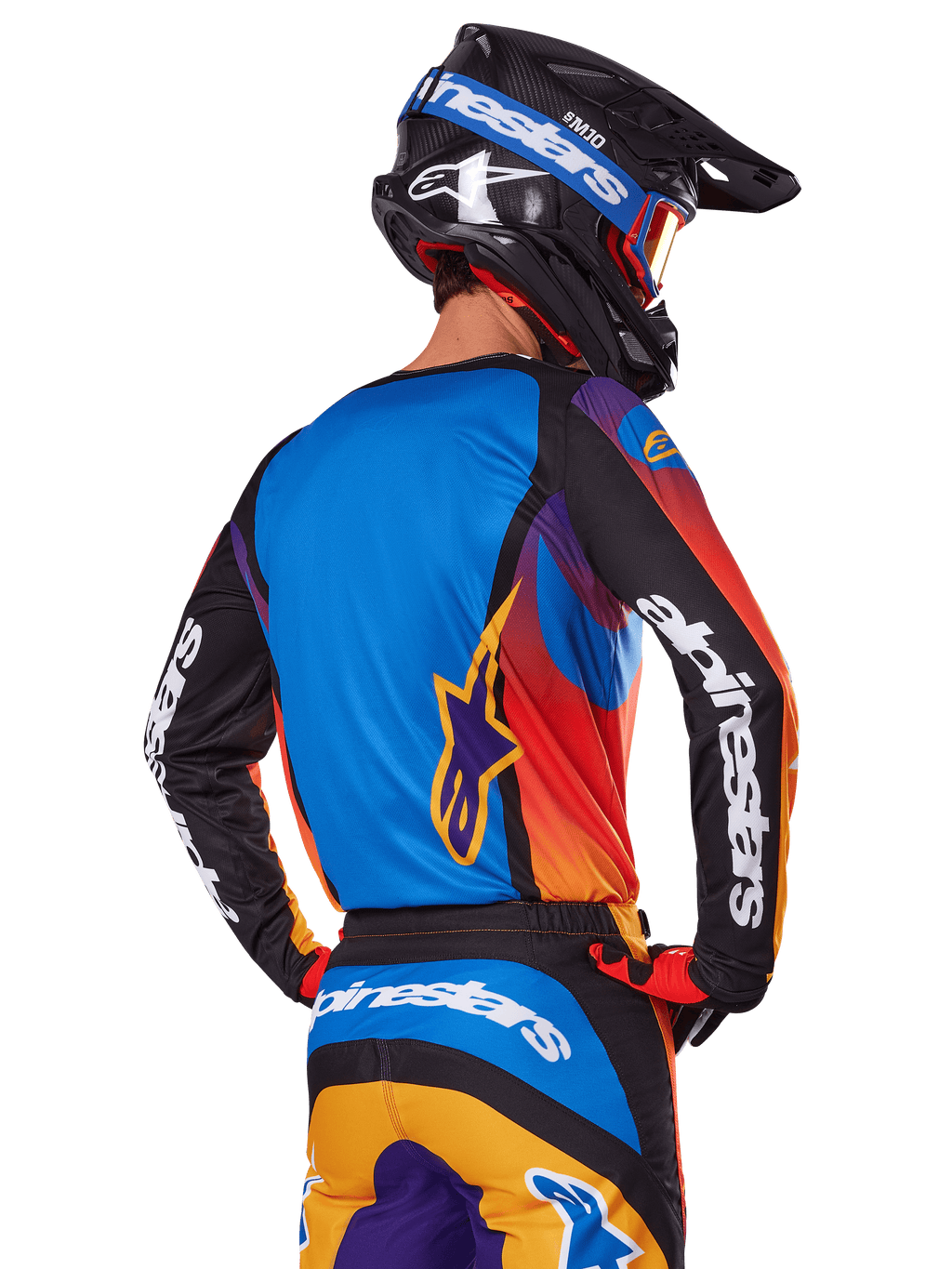 Piloto de motocross visto desde atrás con el conjunto de equipación Alpinestars 2025 Fluid Wurx, jersey multicolor con espalda azul y mangas negras con logotipos blancos, pantalones a juego en naranja y azul, casco negro y azul con gafas naranjas, atuendo de carreras off-road coordinado.