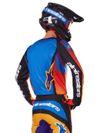 Piloto de motocross visto desde atrás con el conjunto de equipación Alpinestars 2025 Fluid Wurx, jersey multicolor con espalda azul y mangas negras con logotipos blancos, pantalones a juego en naranja y azul, casco negro y azul con gafas naranjas, atuendo de carreras off-road coordinado.