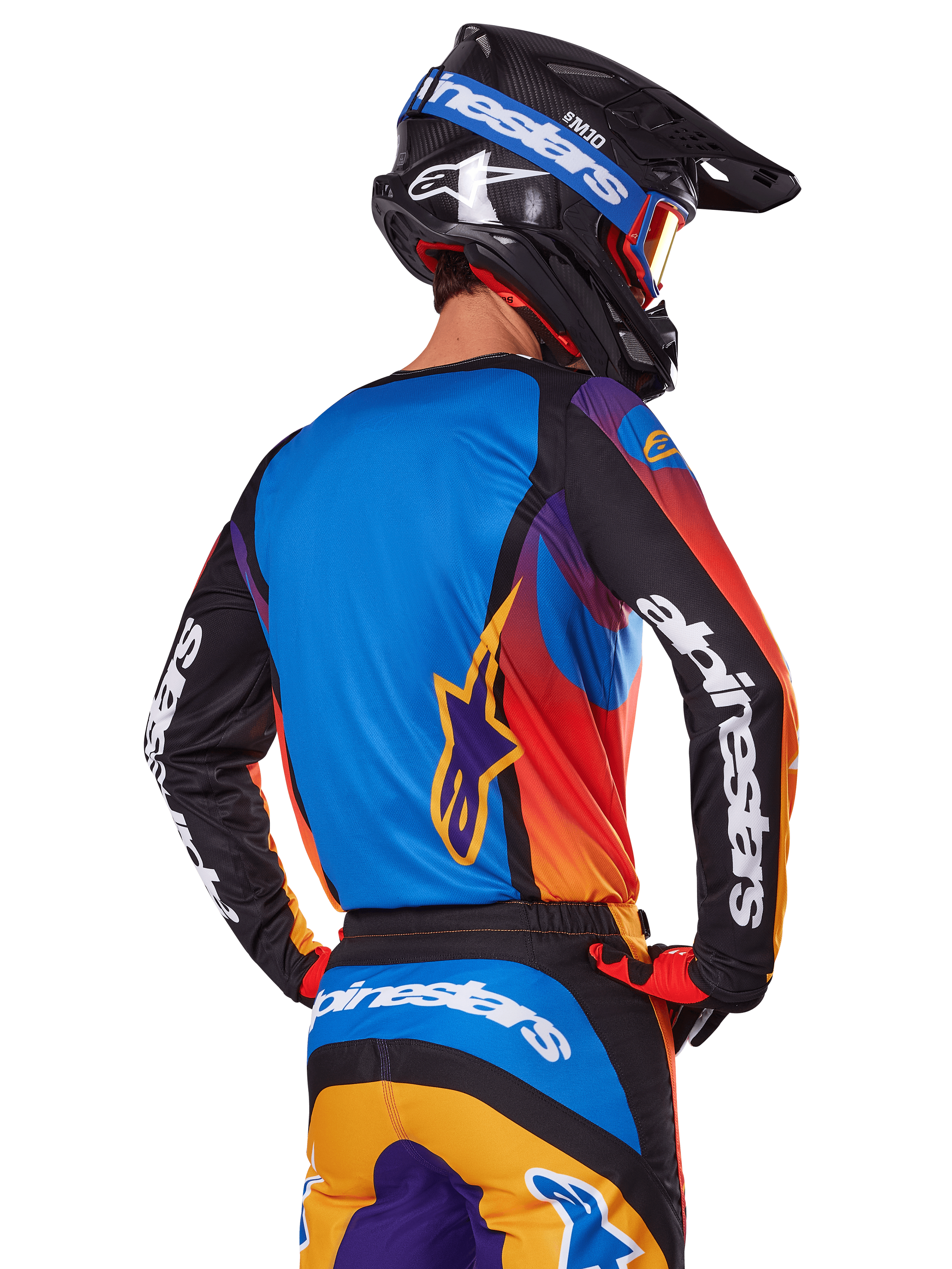Piloto de motocross visto desde atrás con el conjunto de equipación Alpinestars 2025 Fluid Wurx, jersey multicolor con espalda azul y mangas negras con logotipos blancos, pantalones a juego en naranja y azul, casco negro y azul con gafas naranjas, atuendo de carreras off-road coordinado.