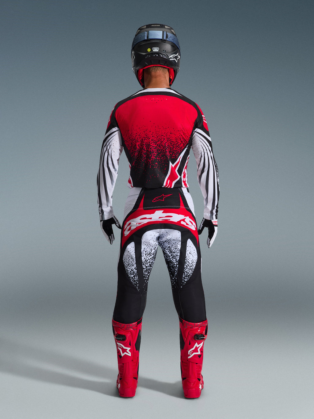 Piloto de motocross visto desde atrás con el conjunto Alpinestars 2026 Techstar Nomur, jersey rojo y blanco con degradado moteado negro, pantalones blancos y rojos a juego con detalles negros, botas de color rojo brillante y casco negro, equipamiento de carreras off-road coordinado.