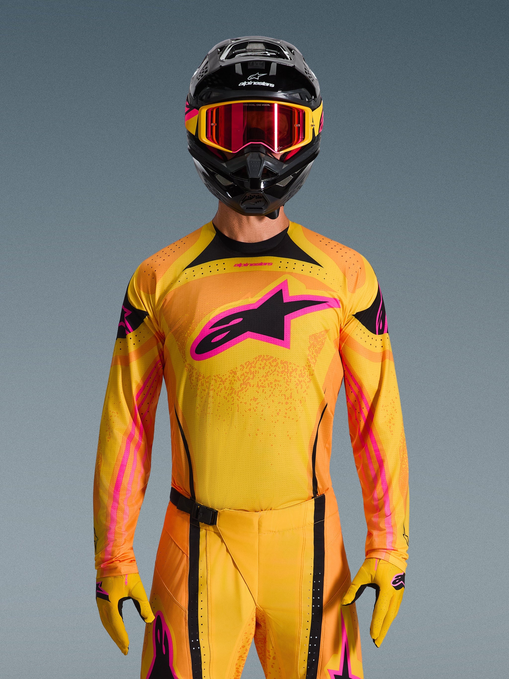 Alpinestars Techstar Nomur Jersey 2026 en naranja, amarillo y rosa, lucido por un piloto de motocross con casco negro y gafas rosas a juego, con un gran logotipo rosa de Alpinestars en el pecho, ajuste atlético de competición para rendimiento off-road.