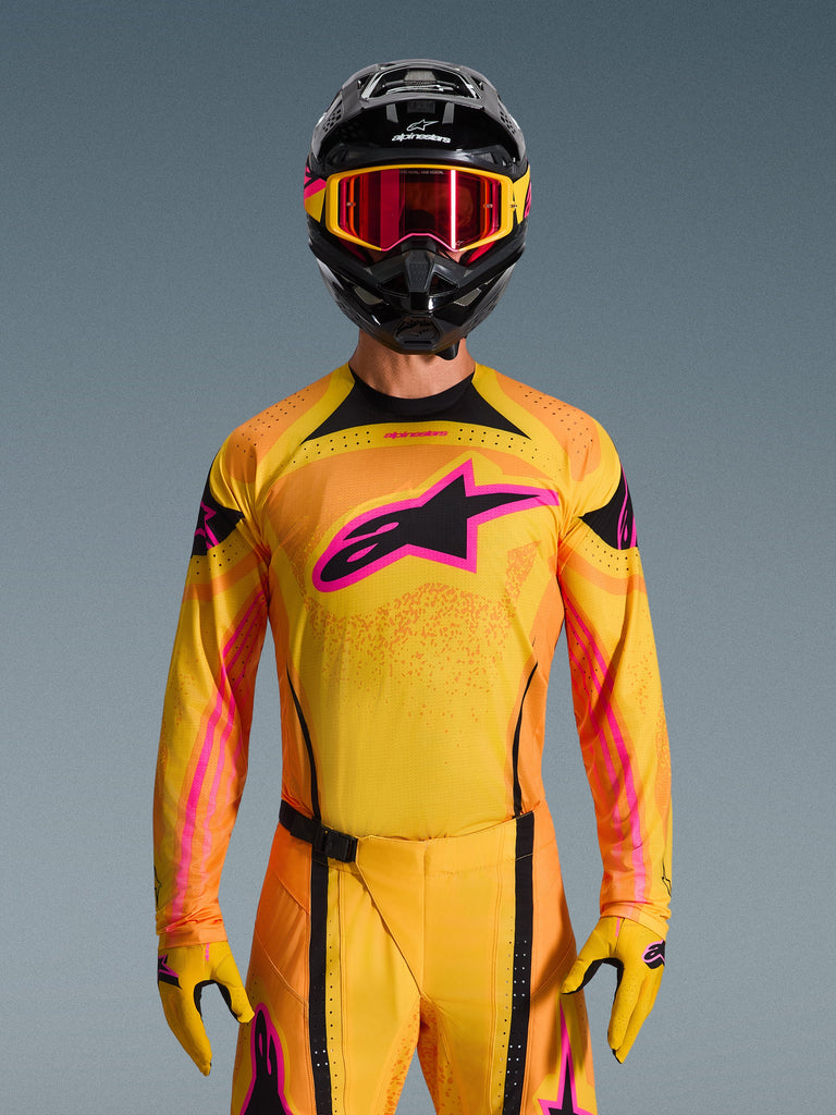 Alpinestars Techstar Nomur Jersey 2026 en naranja, amarillo y rosa, lucido por un piloto de motocross con casco negro y gafas rosas a juego, con un gran logotipo rosa de Alpinestars en el pecho, ajuste atlético de competición para rendimiento off-road.