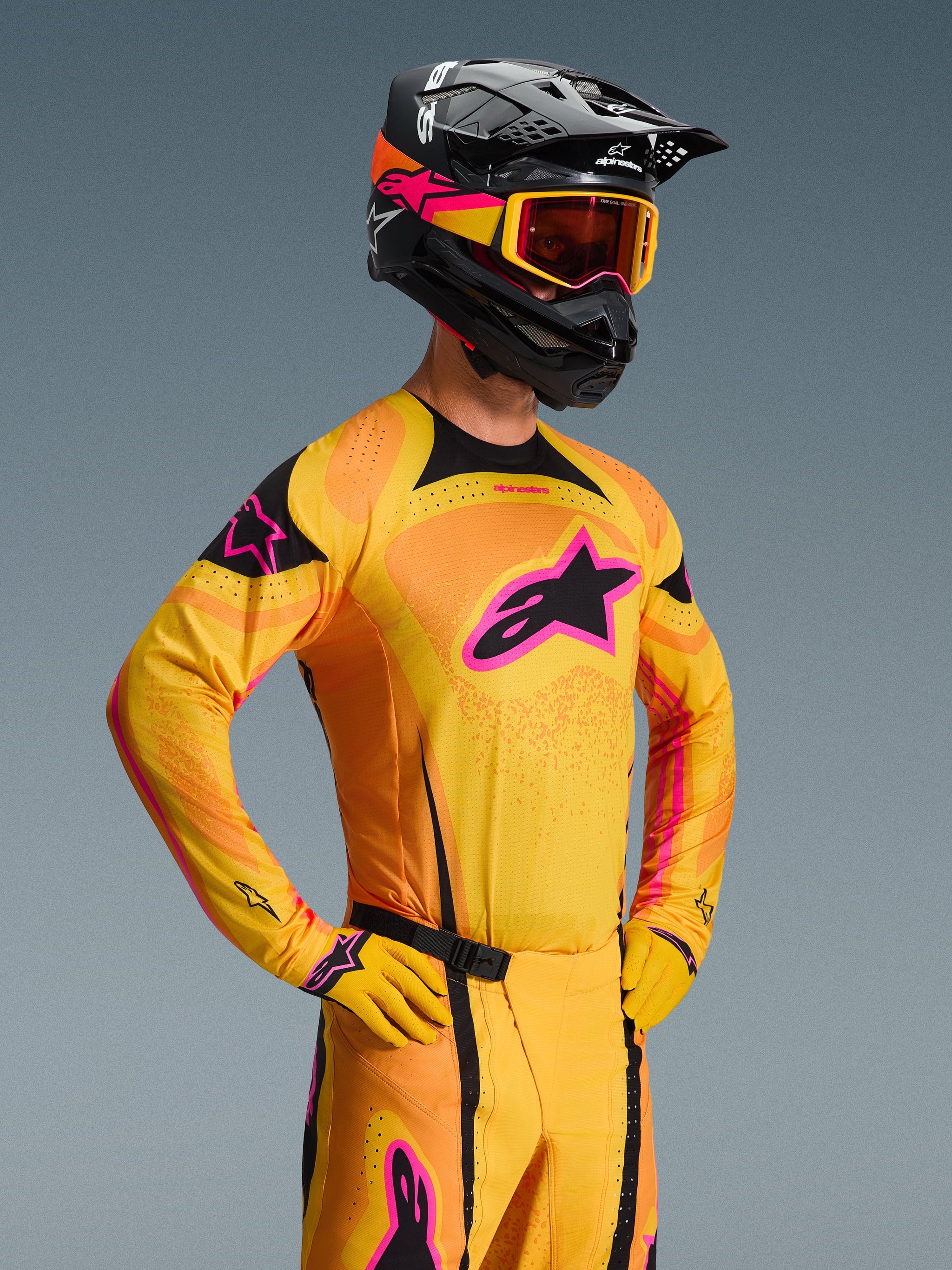 Alpinestars 2026 Techstar Nomur Jersey, camiseta de motocross, naranja, amarillo y rosa, usada por un piloto con casco y gafas negras a juego, con un gran logo de estrella rosa en el pecho, detalles en negro y detalles de ventilación cortados con láser.