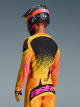Alpinestars Techstar Nomur Jersey 2026 en naranja, amarillo y rosa, vista posterior de un piloto de motocross con equipamiento completo que incluye pantalones a juego y casco negro, diseño vibrante con detalles en amarillo y rosa y panel trasero negro, ajuste de carrera atlético para rendimiento off-road