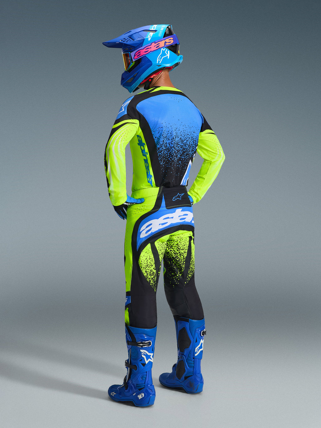 Motocross rider viewed from behind wearing the Alpinestars 2026 Techstar Nomur Gear Set, featuring a fluorescent yellow and blue jersey with black speckle accents, matching pants with bold white branding, blue racing boots, and a blue helmet, complete professional off-road racing outfit