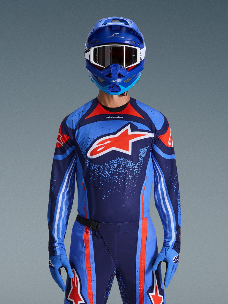 Piloto de motocross con el jersey Alpinestars Techstar Nomur 2026 en azul marino oscuro, naranja y azul UCLA, pantalones a juego y guantes azules, casco azul con gafas blancas, logotipo grande de Alpinestars en rojo y blanco en el pecho, equipamiento de rendimiento coordinado para carreras todoterreno.