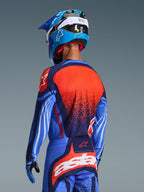 Motocross rider from the back wearing the Alpinestars 2026 Techstar Nomur Jersey and matching pants, Dark Navy and UCLA Blue with vibrant orange accents, featuring a large star graphic and laser-cut ventilation on the jersey, paired with a blue and white helmet for a complete off-road racing kit