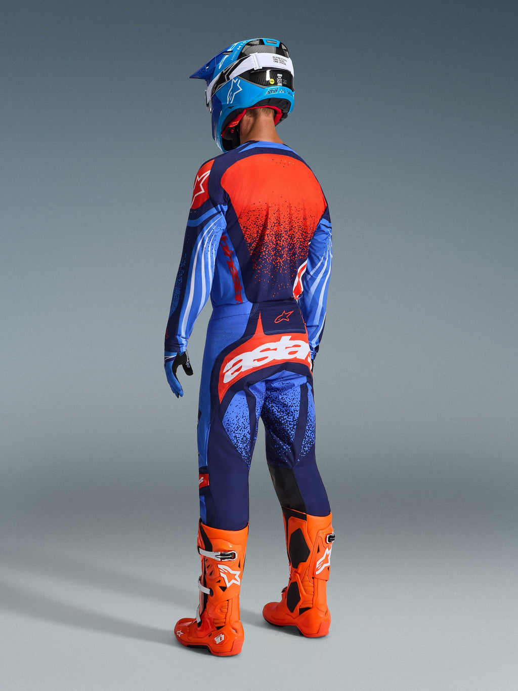 Motocross rider from the back wearing the 2026 Techstar Nomur Gear Set, Dark Navy and UCLA Blue jersey with orange panel, matching pants with orange 'astars' logo, bright orange motocross boots, and a blue and white helmet, coordinated competition racing gear