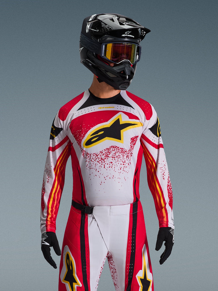 Alpinestars Techstar Nomur Jersey 2026, jersey de motocross, rojo, blanco y negro, con un gran logotipo negro de la estrella A en el pecho con detalles amarillos, diseño de salpicaduras rojas, usado con un casco negro y equipamiento off-road a juego.