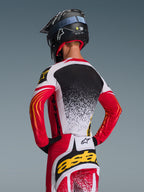Alpinestars 2026 Techstar Nomur Gear Set, Motocross Jersey and Pants, Red White and Black with yellow accents, back view of rider wearing a black helmet, performance racing apparel with splatter graphics and bold branding