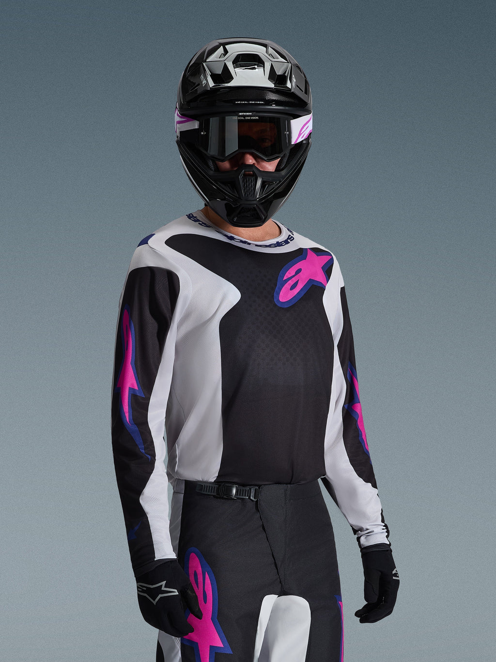 Piloto de motocross con el Jersey Alpinestars Fluid Grid 2026 en negro, gris claro y púrpura, con grandes logotipos de estrellas en rosa y púrpura, combinado con pantalones a juego, casco negro y gafas, conjunto completo de equipamiento para carreras off-road