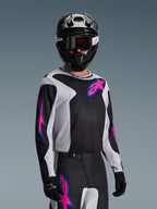 Piloto de motocross con el Jersey Alpinestars Fluid Grid 2026 en negro, gris claro y púrpura, con grandes logotipos de estrellas en rosa y púrpura, combinado con pantalones a juego, casco negro y gafas, conjunto completo de equipamiento para carreras off-road