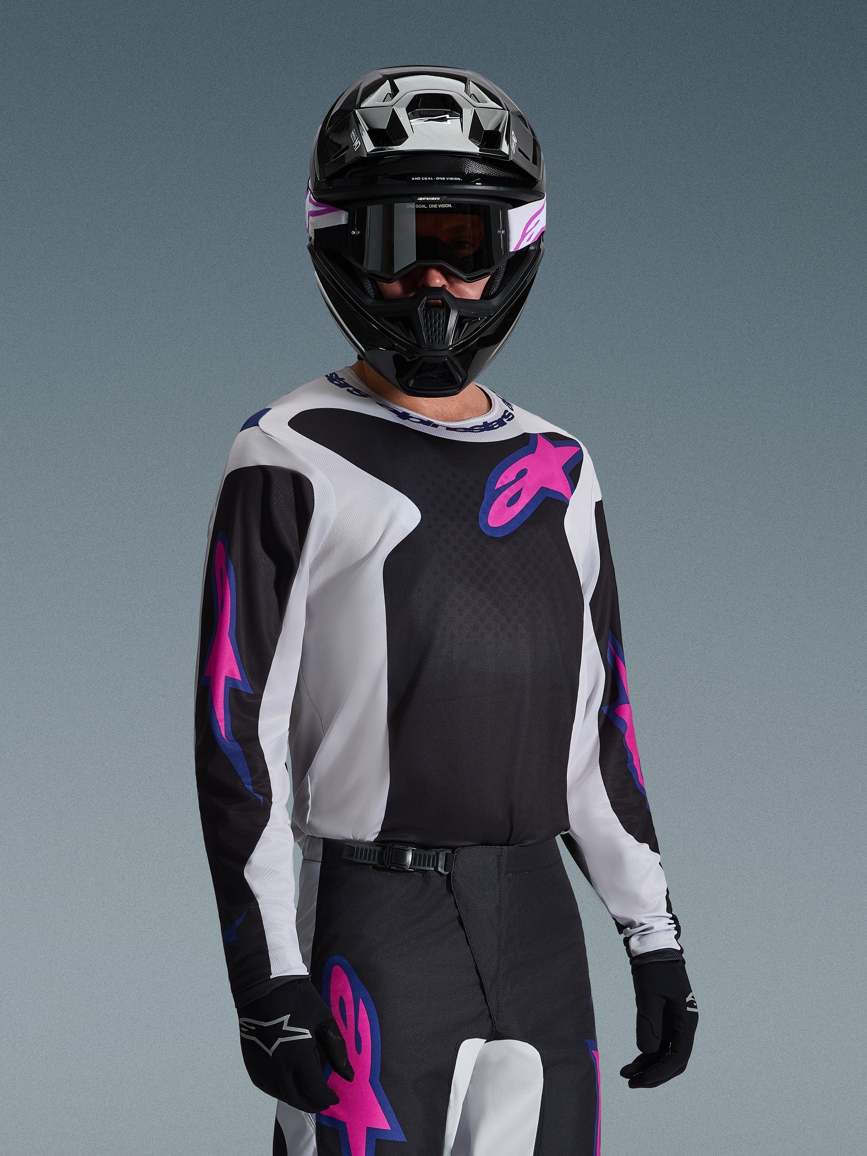Piloto de motocross con el Jersey Alpinestars Fluid Grid 2026 en negro, gris claro y púrpura, con grandes logotipos de estrellas en rosa y púrpura, combinado con pantalones a juego, casco negro y gafas, conjunto completo de equipamiento para carreras off-road