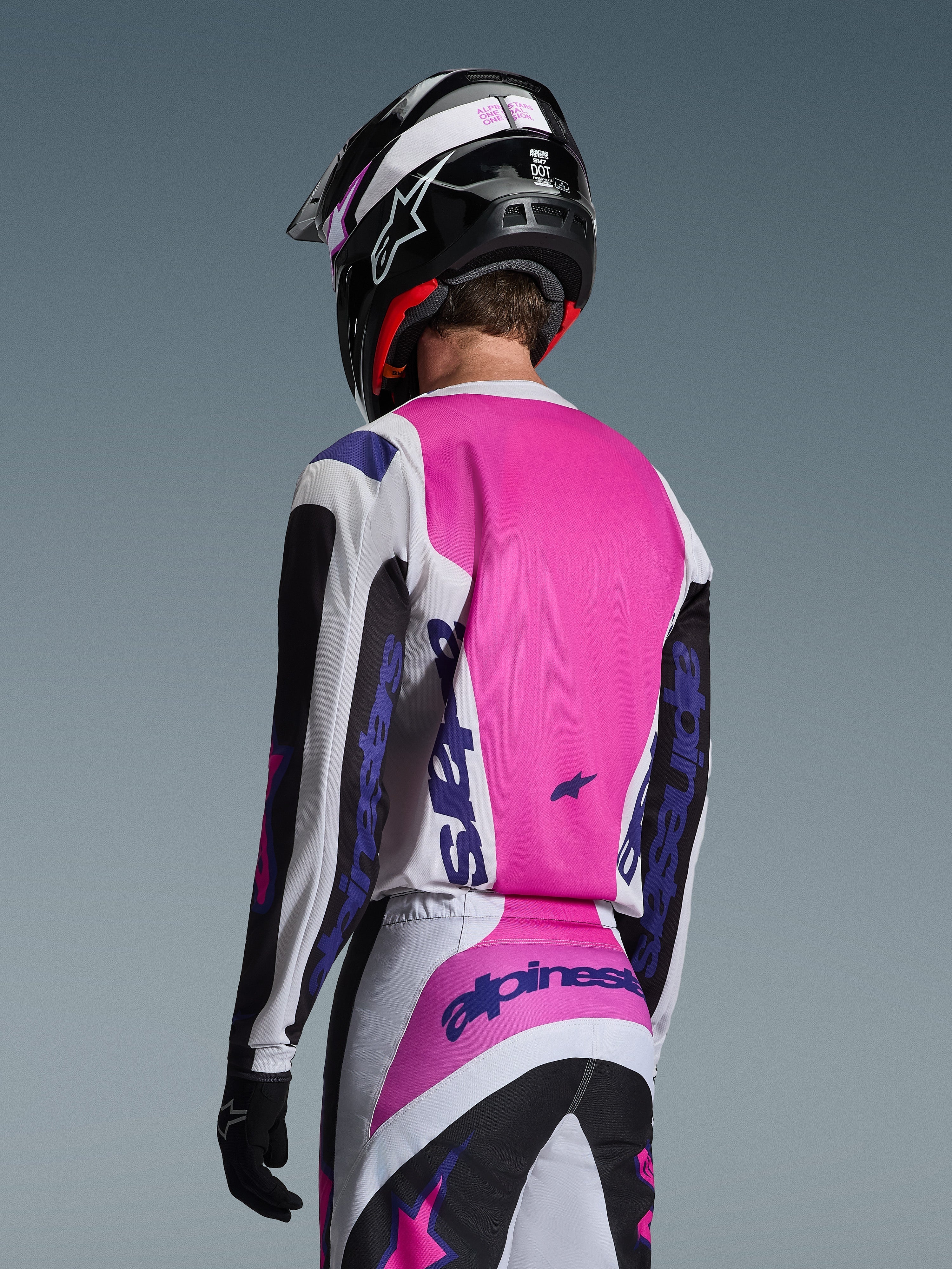 Piloto de motocross visto desde atrás vistiendo el Jersey Alpinestars Fluid Grid 2026 en negro, blanco y púrpura, pantalones a juego con logotipos llamativos, casco negro con detalles en rosa y guantes negros; equipamiento completo coordinado para el rendimiento off-road.